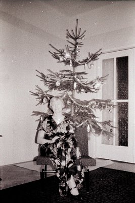 Vintage photograph shows a seated woman in a patterned dress next to a sparsely decorated, artificial Christmas tree. Interio...