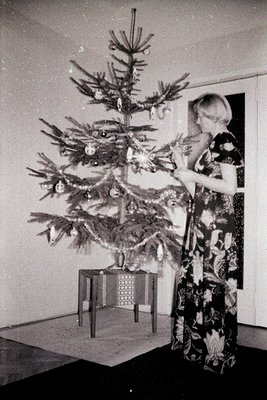A young woman in a floral dress decorates a sparsely adorned Christmas tree. The scene captures a mid-century modern aestheti...
