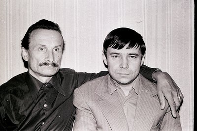 Two men stand close, posed formally. The older man, with a prominent mustache, has an arm around the younger man’s shoulder. ...
