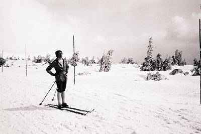 A man stands on skis in a snow-covered alpine landscape. He wears a tailored suit, knickerbockers, and holds ski poles. Spars...