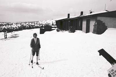 A person in a suit and skis stands outdoors before a simple, one-story building with a steep, gabled roof. Snowy landscape ex...