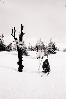 A lone skier rests near a weathered signpost and small shelter on a snowy, mountainous landscape. Appears to be a recreationa...