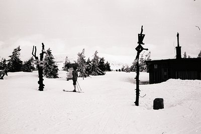 A lone skier pauses on a snow-covered slope beside a rustic wooden building, likely a mountain lodge. Signposts point in vari...