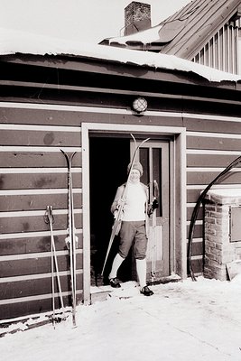 A man stands in a doorway of a distinctive, horizontally-lined wooden structure, holding vintage skis. He wears a knit cap, s...