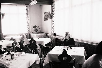 A monochrome image captures an elderly gentleman seated at a table within what appears to be a traditional restaurant or dini...