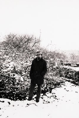 Man stands in snowy landscape, bundled in overcoat and hat. Bare trees with snow-laden branches frame the scene. Likely mid-2...