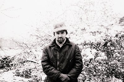 Man in a dark coat & hat stands amidst snow-laden shrubbery, appearing contemplative. Visible details include a thick collar ...