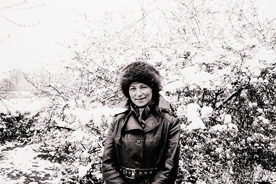 A woman stands bundled in a fur-trimmed coat and hat, posed against a snow-laden landscape. The scene conveys a sense of cold...