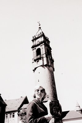Tall, monumental clock tower dominates this image, rising above a woman in a dark suit and spectacles. Likely a European sett...