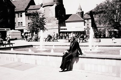 Man in a suit sits on a stone ledge overlooking a tiered fountain in a town square. Buildings with ornate facades line the pe...