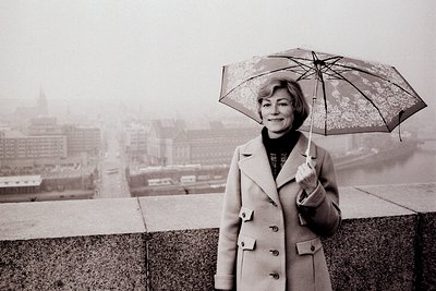 A woman stands on a raised parapet overlooking a city shrouded in mist. She wears a double-breasted coat and holds an ornate,...