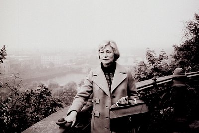 A woman in a wool coat stands on a stone parapet overlooking a hazy cityscape with a river and distant buildings. She carries...