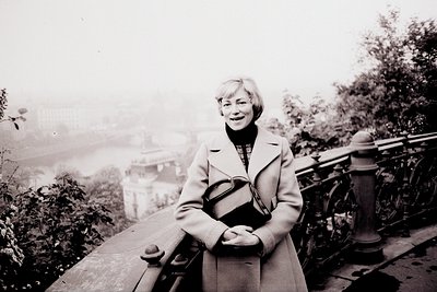 A smiling woman in a belted coat & turtleneck stands on a stone parapet overlooking a city shrouded in mist. She carries a ba...