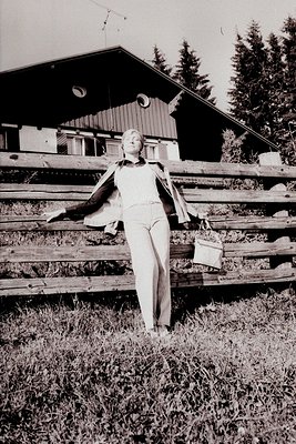 A woman in wide-leg trousers and a patterned top leans against a wooden fence. A timber-framed building with a satellite dish...
