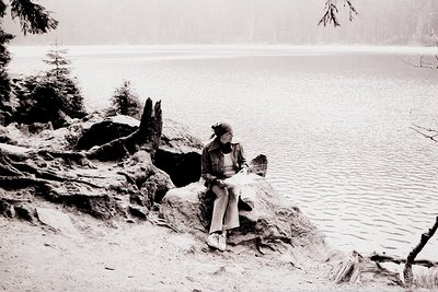 A lone woman sits on a rocky shoreline overlooking a tranquil lake. She wears a turtleneck sweater, a jacket, and a headscarf...