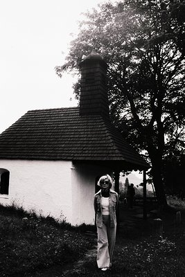 A woman in flared pants and a vest stands before a small, whitewashed building with a steep, shingled roof and tall chimney. ...