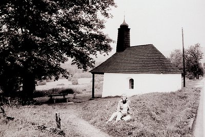 Quaint roadside shrine, likely Eastern European, features a steeply pitched roof & small window. A seated woman in vintage cl...