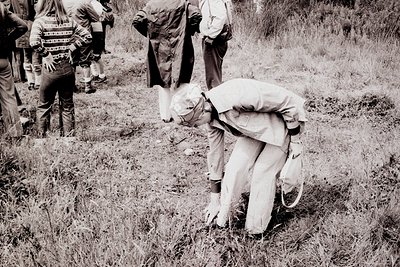 A man in a suit and tie bends over, inspecting the ground. Two figures in casual clothing stand behind him. The photo, likely...