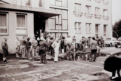A group gathers outside a stark, modernist building - likely a government or industrial structure. Men in wide-legged trouser...
