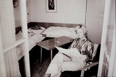 Woman in chair reads newspaper in sparsely furnished room. 1970s style decor with simple bed, small side table, and patterned...