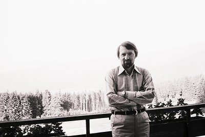 Man in a striped shirt and belted trousers stands with arms crossed on a balcony overlooking a snow-covered, forested landsca...