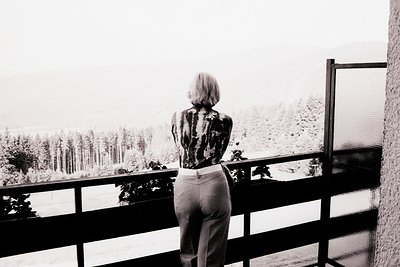 A person with short, cropped hair stands on a balcony overlooking a dense, snow-covered forest. The subject wears a patterned...