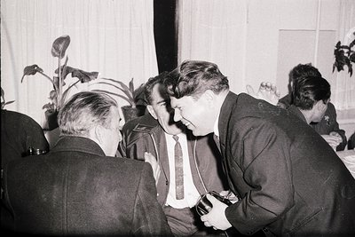 Three men in formal wear engage in conversation within a warmly lit, indoor setting. A fourth man, partially visible, stands ...