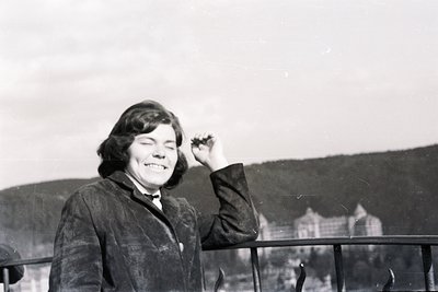 A smiling young woman in a dark jacket stands behind a balcony railing, arm raised. A building sits on a hillside backdrop. A...