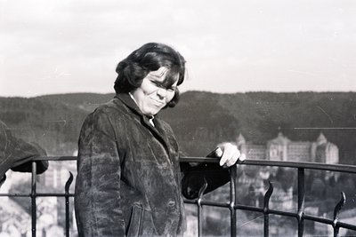 A woman in a dark, textured coat stands at a wrought-iron railing, overlooking a town with distinctive architecture, possibly...