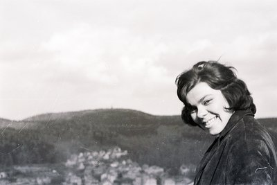 A smiling young woman with a short, dark hairstyle is posed outdoors. She wears a dark, collared jacket and gazes towards the...