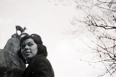 A woman in a dark coat gazes slightly off-camera. A bronze statue of a dog stands atop a stone base behind her. Stark tree br...