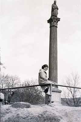 A young woman in a 1960s-style coat sits playfully on a stone pedestal topped with a statue. The neoclassical monument stands...