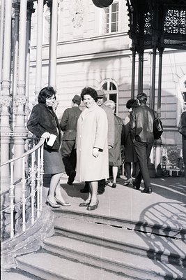 Elegant monochrome photo captures a 1960s or 70s European scene, likely a seaside resort. A group of formally dressed people—...