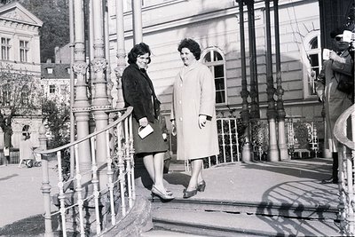 Two women in 1960s attire chat on a stone staircase with ornate iron railings and a grand building backdrop. One wears a dark...