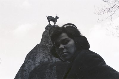 A person with dark hair and a jacket looks directly at the camera, positioned at the base of a rocky outcrop. A mountain goat...