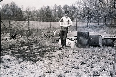A shirtless man stands in a muddy garden, hands on hips, next to stacked barrels and gardening tools. A chicken pecks in the ...