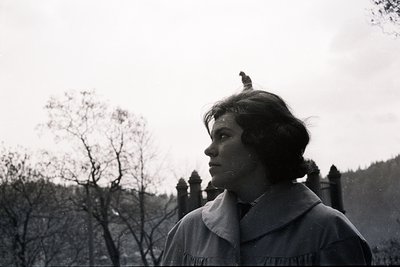 A woman in a high-collared coat gazes to the side, captured in a grainy, black and white outdoor portrait. Sparse tree branch...