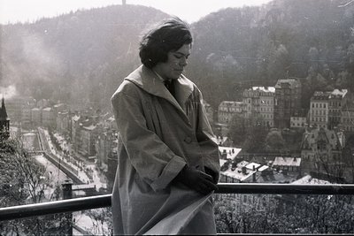 A woman in a coat stands at a balcony overlooking a city nestled in a valley. The cityscape shows traditional architecture wi...
