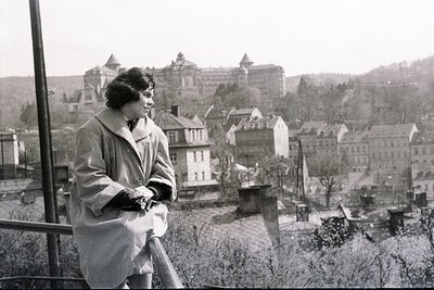 A woman in a tailored coat stands poised on a balcony overlooking a hillside town. The architecture suggests a European setti...