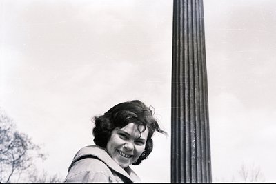 A smiling woman in a raincoat stands near a tall, fluted column. Appears to be a candid portrait, possibly taken in the 1960s...