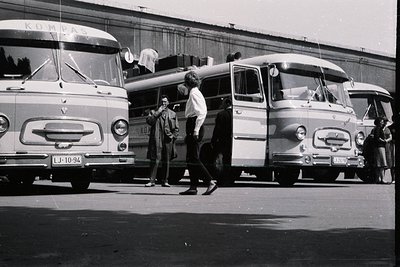 Several "Kompas" buses, likely used for tourism, line a curb. People in 1960s attire wait nearby. The buses display "LJ-10-94...