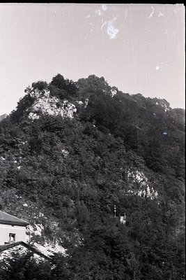 Dense alpine forest blankets a steep hillside, revealing rocky outcrops. A small structure, possibly a chapel or lookout, sit...