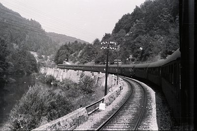 A long passenger train curves along a railway track built beside a river, flanked by dense, wooded hills. Stone retaining wal...