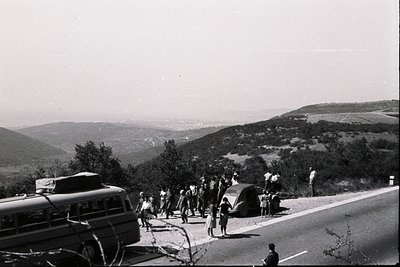 A group of people disembarks from a vintage bus alongside a classic convertible car, set against a backdrop of rolling hills ...