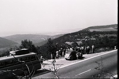 A group gathers beside a vintage bus and car on a hillside road. A distant town sits in a valley below. Likely a tourist stop...