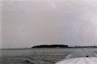 A low-angle, monochrome view captures a coastal landscape. A small island with dense vegetation sits offshore, framed by the ...