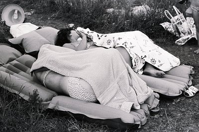Woman reclines on inflatable pillows and a mattress, covered with a patterned sheet. Seaside setting, likely a beach or grass...