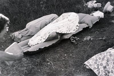 Two individuals recline on an inflatable mattress within a grassy, outdoor setting. One wears a patterned dress and sandals, ...