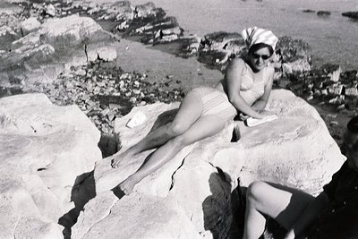 A young woman reclines on rocks, reading a book while wearing a vintage swimsuit and headscarf. Likely a vacation snapshot fr...