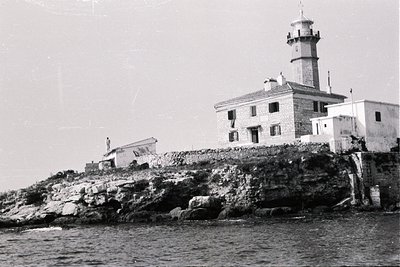 Stone lighthouse complex sits atop a rocky promontory, overlooking the sea. A small figure is visible near a low, attached st...
